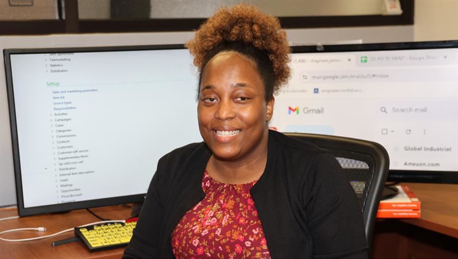 A smiling woman with curly hair sits at a desk with 2 monitors, a yellow keyboard, & books. 